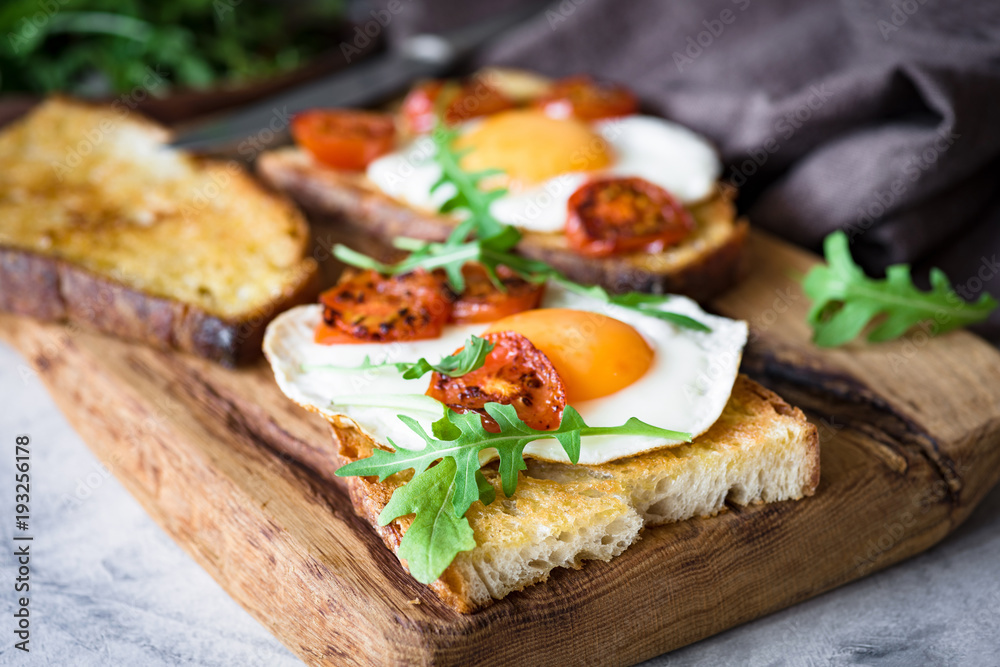 Healthy breakfast toast with egg, roasted tomato and arugula on rustic wooden cutting board. Closeup view