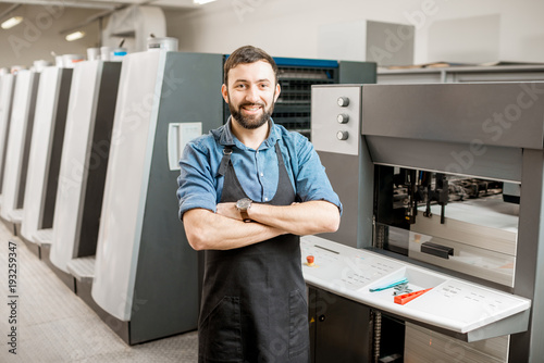 Portrait of a handsome typographer standing at the printing manufacturing with offset machine on the backgorund