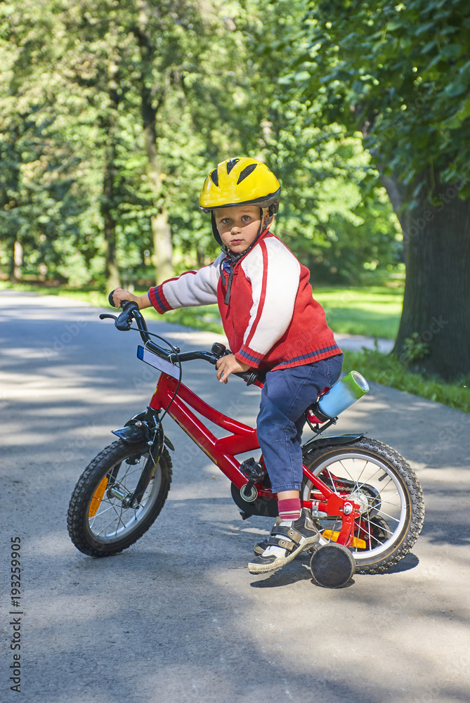 Little boy on bicycle in green park outdoor in summer. A child is riding a children's bike with support wheels  wearing safety helmet