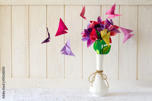 Origami paper flowers in vase and butterflies on white wooden wall. Paper art craft