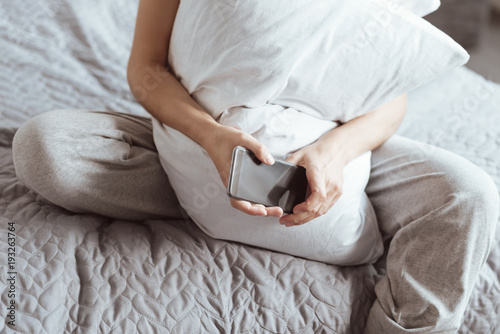 Cozy home evening. Top view on a young lady wearing pajamas sitting on a bed and embracing a pillow while holding her phone.