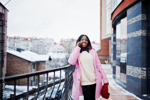 African american girl in red hat and pink coat at street of city against building on winter day with mobile phone. © AS Photo Family