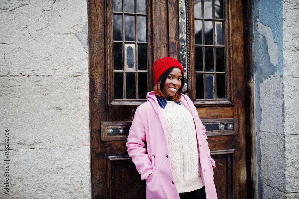African american girl in red hat and pink coat against wooden door at street of city. African american girl in red hat and pink coat against wooden door at street of city.