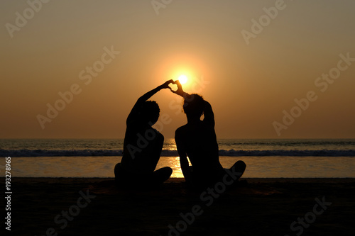Couple's silhouette drawing heart with their hands on the beach at sunset