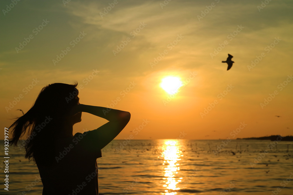 Silhouette of young woman watching the sun rising on golden sky with a flying bird 