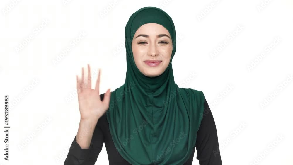 Portrait of friendly oriental woman in traditional arabic scarf welcoming and greeting with waving hand and smile, over white background. Concept of emotions Portrait of friendly oriental woman in traditional arabic scarf welcoming and greeting with waving hand and smile, over white background. Concept of emotions