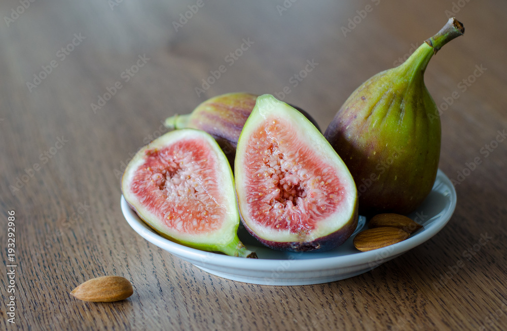 Close-up of fresh figs with one slised ripe fig and three almond nuts lying on white plate on a wooden table background Close-up of fresh figs with one slised ripe fig and three almond nuts lying on white plate on a wooden table background