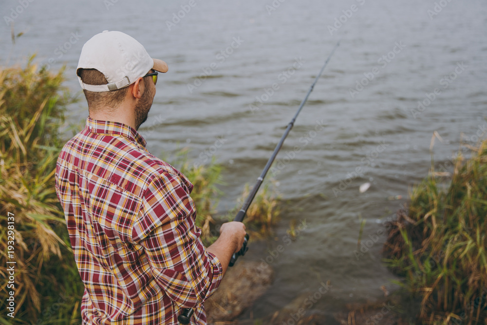Back view Young unshaven man with a fishing rod in checkered shirt, cap, sunglasses casts bait and fishing on lake from shore near shrubs and reeds. Lifestyle, recreation, fisherman leisure concept Back view Young unshaven man with a fishing rod in checkered shirt, cap, sunglasses casts bait and fishing on lake from shore near shrubs and reeds. Lifestyle, recreation, fisherman leisure concept