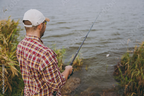 Back view Young unshaven man with a fishing rod in checkered shirt, cap, sunglasses casts bait and fishing on lake from shore near shrubs and reeds. Lifestyle, recreation, fisherman leisure concept