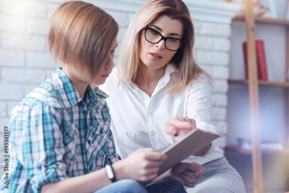 Fill the blank space. Serious woman pointing at a clipboard held by a teenage boy passing a test while attending a professional psychologist. Fill the blank space. Serious woman pointing at a clipboard held by a teenage boy passing a test while attending a professional psychologist.