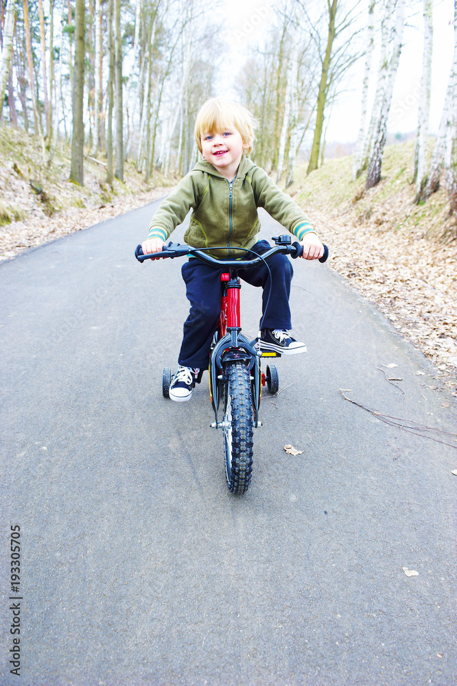 Little boy on bicycle in green park outdoor in summer. A child is riding a children's bike with support wheels wearing safety helmet Little boy on bicycle in green park outdoor in summer. A child is riding a children's bike with support wheels wearing safety helmet