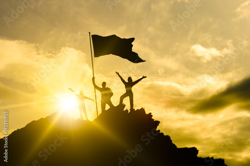 High success, family three silhouette, father of mother and child holding flag of victory on top of mountain, hands up. A man on top of a mountain. Against the dramatic sky at sunset.