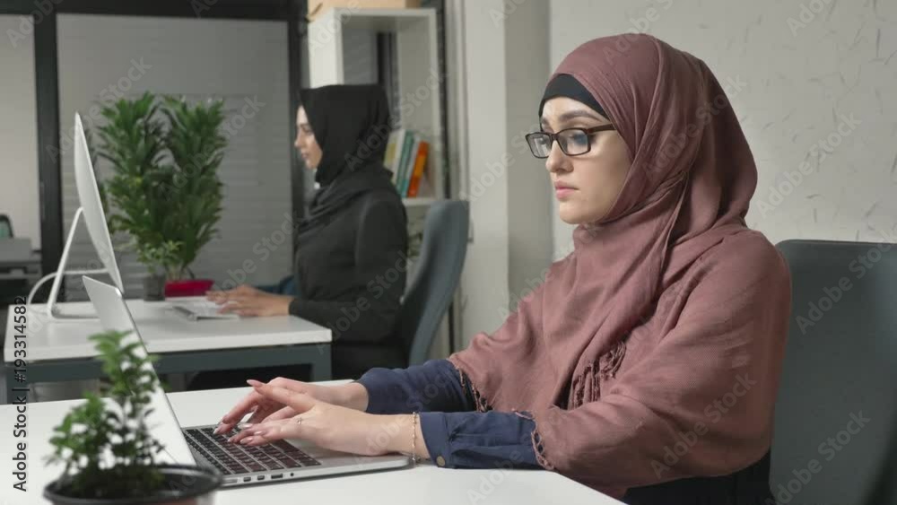 Young beautiful girl in pink hijab and glasses typing, working on computer. Look at the camera. Arab women in the office. Girl in black hijab in background. 60 fps Young beautiful girl in pink hijab and glasses typing, working on computer. Look at the camera. Arab women in the office. Girl in black hijab in background. 60 fps