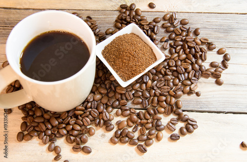 A cup of coffee with a small white ceramic dish full of ground coffee and coffee beans on wooden background.