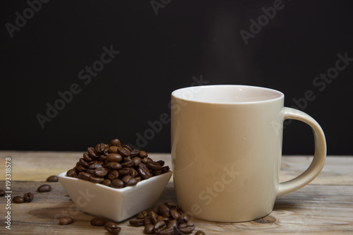 A cup of coffee with small white ceramic dish full of coffee beans on wooden bakcground.