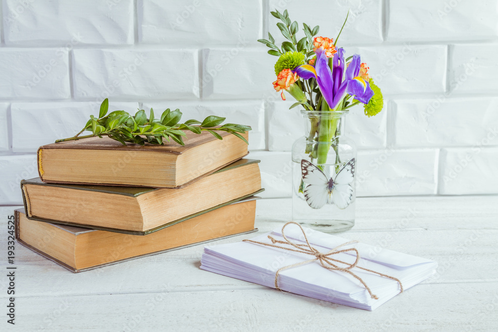 Old books and envelopes, a vase of flowers against a white brick wall.