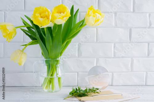 On a background of a white brick wall a vase with yellow tulips, an envelope and a bottle of perfume.