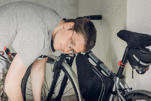 Young blond man with short haircut in gray t-shirt with glasses bending over his sports bicycle