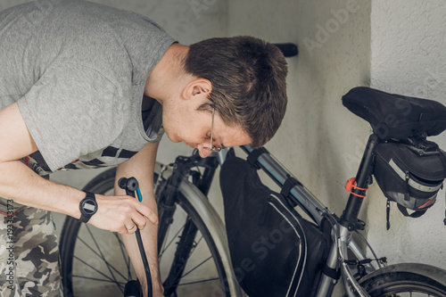 Young blond grey-eyed man with short haircut in gray t-shirt with glasses bending over his sports bike and holding bicycle pump