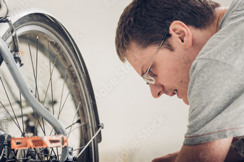 Young blond man with short haircut in gray t-shirt with glasses looking carefully at the wheel of broken bicycle