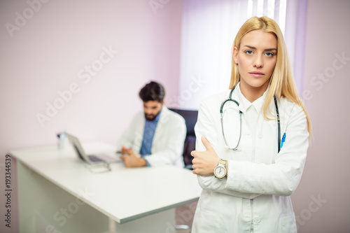 Portrait of blonde serious female doctor while standing in front of camera  male doctor in background working on the computer.