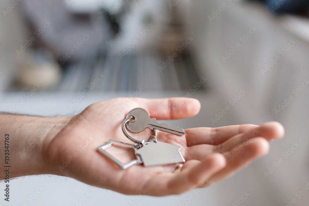 Men hand holding key with house shaped keychain. Modern light lobby interior. Mortgage concept. Real estate, moving home or renting property. Men hand holding key with house shaped keychain. Modern light lobby interior. Mortgage concept. Real estate, moving home or renting property.