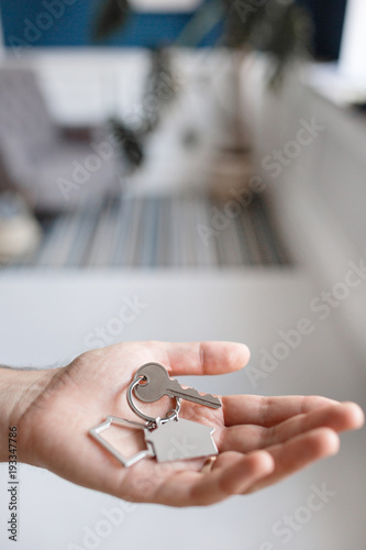 Men hand holding key with house shaped keychain. Modern light lobby interior. Mortgage concept. Real estate, moving home or renting property.