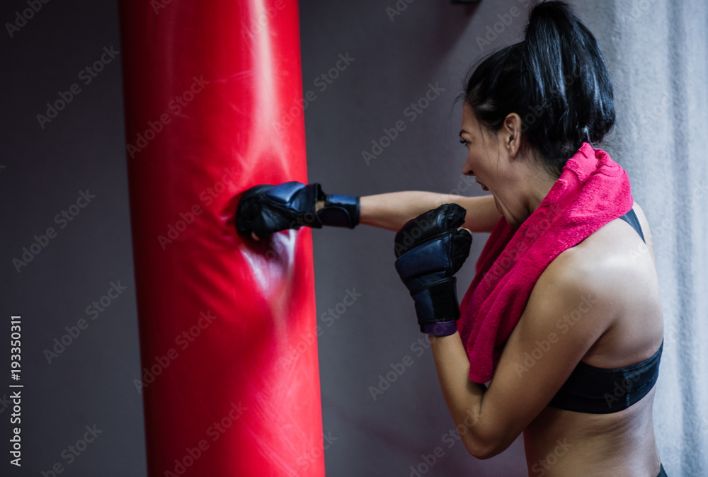 Horizontal back view of athlete woman with red towel on neck punching the red bag in kickboxing gloves at the gym. Woman boxer workout. Sport, fitness, lifestyle, people and motivation concept.