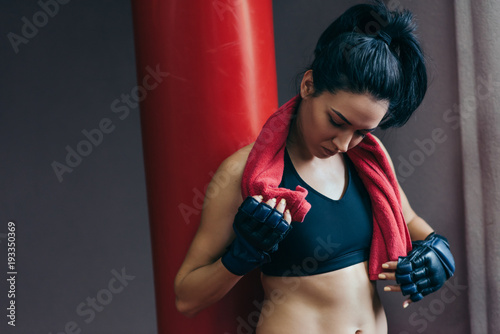 Caucasian attractive brunette sportswoman with red towel on neck before workout with punch bag and wearing kickboxing gloves waiting trainer in the gym. Sport, fitness, people concept.
