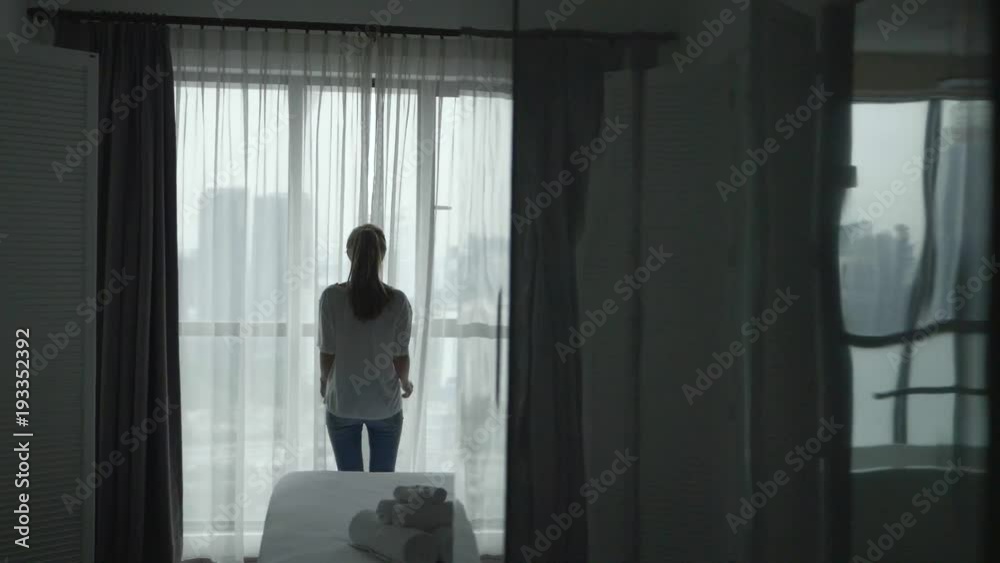 Young woman uncover the big window and looking out her apartment on the city buildings. Young girl unveil curtains in room in the cloudy morning in slow motion Young woman uncover the big window and looking out her apartment on the city buildings. Young girl unveil curtains in room in the cloudy morning in slow motion