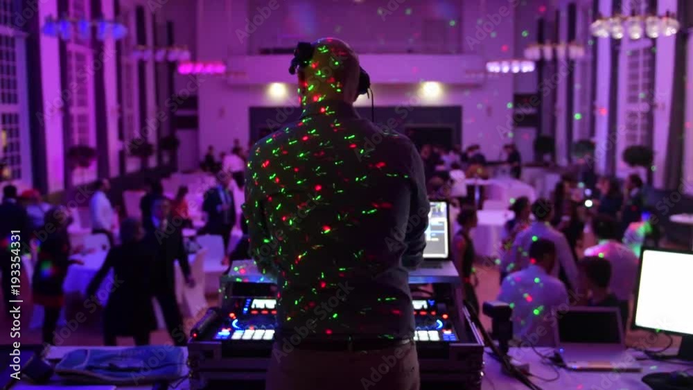 Unrecognizable Music DJ mixing dancing in front his turntables and mixer and laptop during party wedding - illumination with laser light of his back and large room with guests