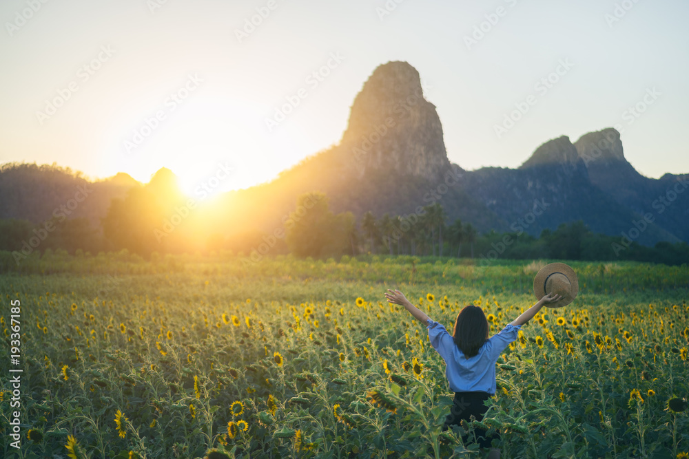 Young woman traveler with hat in sunflower fields with happiness and cheerful at sunset and arms raised up.