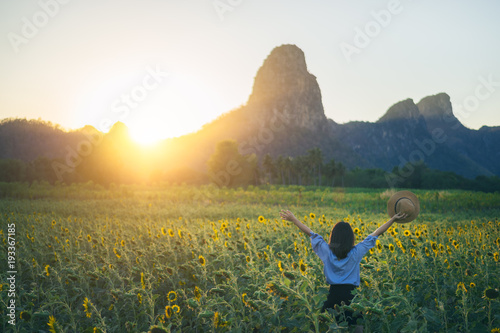 Young woman traveler with hat in sunflower fields with happiness and cheerful at sunset and arms raised up.