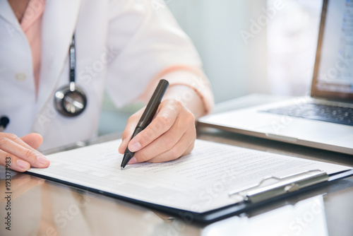 female doctor is using laptop and writing something on clipboard.