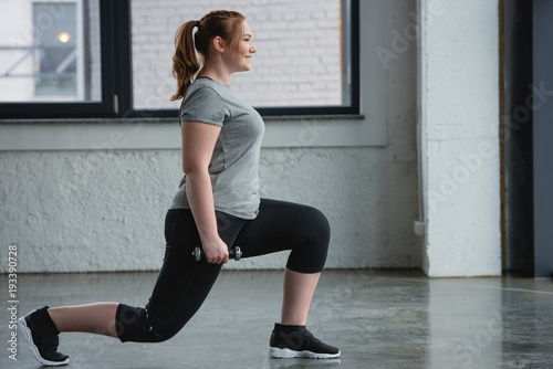Obese girl performing lunges with dumbbell in gym
