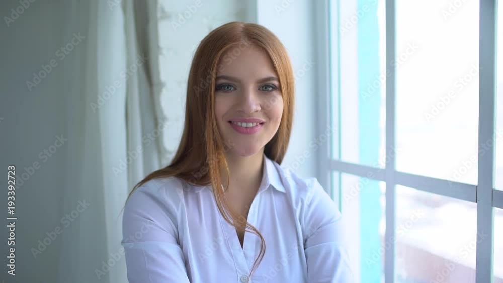 portrait of a smiling redheaded girl sitting at home near a window. slow-motion. portrait of a smiling female student. Woman smiling closeup portrait. Young business woman professional looking at