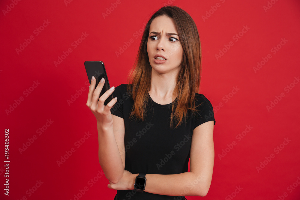 Photo of dissatisfied woman in black clothing looking on mobile phone with confusion or misunderstanding, over red background Photo of dissatisfied woman in black clothing looking on mobile phone with confusion or misunderstanding, over red background