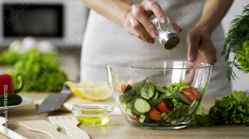 Woman adding salt in vegetable salad glass bowl, health care, excessive salting