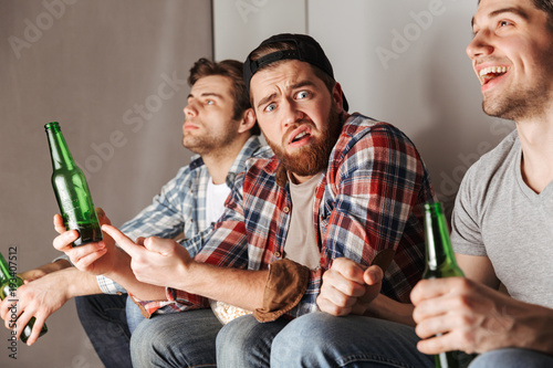 Photo of upset man expressing frustration and dissatisfaction, while watching football game in flat with male friends