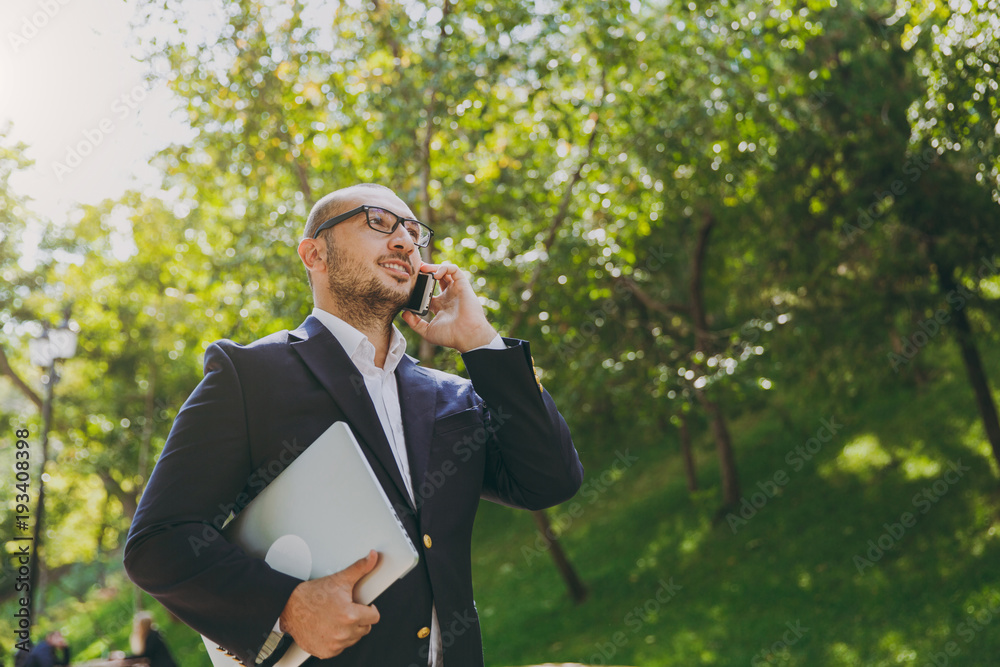 Successful smart businessman in white shirt, classic suit, glasses. Man stand with laptop pc computer, talk on mobile phone in city park outdoors on nature background. Mobile Office, business concept. Successful smart businessman in white shirt, classic suit, glasses. Man stand with laptop pc computer, talk on mobile phone in city park outdoors on nature background. Mobile Office, business concept.
