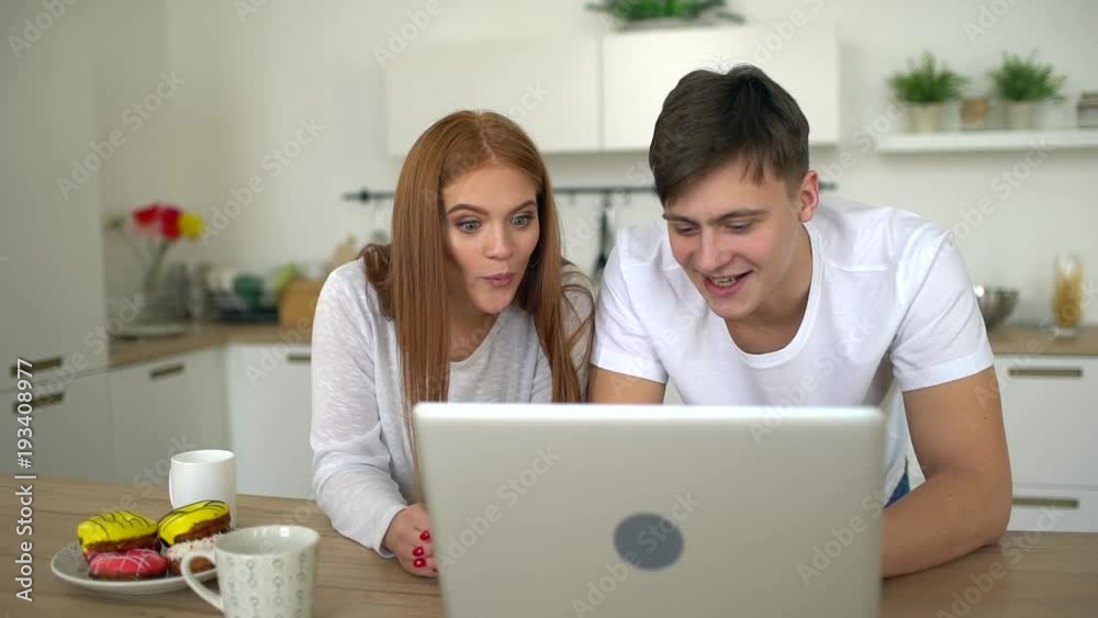 Happy attractive young couple relaxing at home, watching TV or film or foto on laptop. Slow motion. Caucasian couple man and woman is using computer at kitchen. Home, technology and relationships Happy attractive young couple relaxing at home, watching TV or film or foto on laptop. Slow motion. Caucasian couple man and woman is using computer at kitchen. Home, technology and relationships