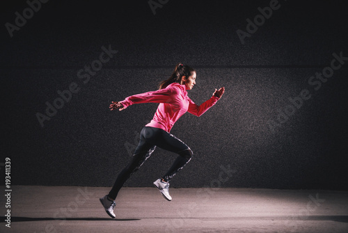 Side motion view of attractive sporty athlete running girl in sportswear at the street in front of the grey wall.
