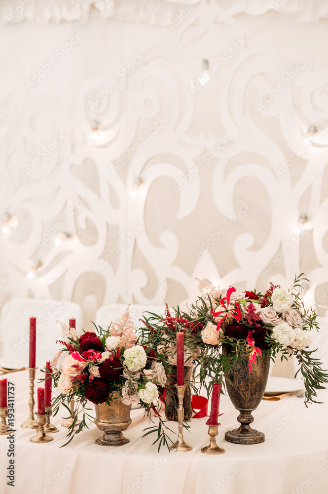 Fototapeta premium Decorate table with white tablecloth, red bouquets with fern and eucalyptus in brass vases, red candles near
