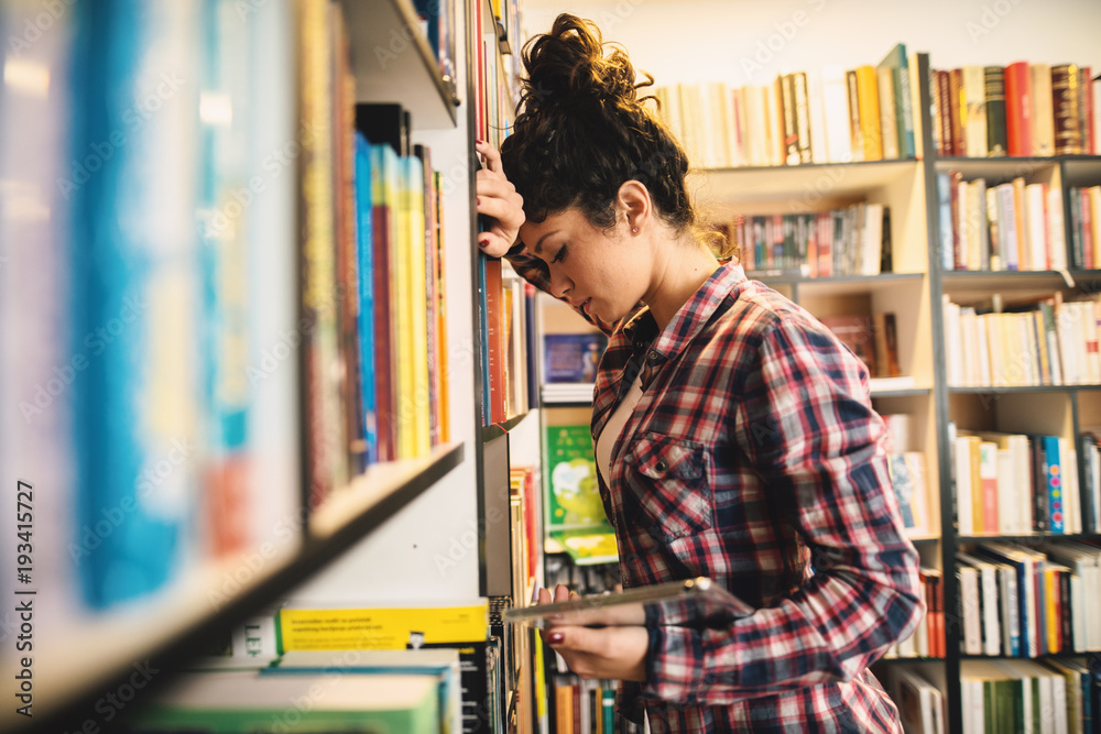 Adorable student girl leaned on the bookshelf in the library looking concerned about the grades and holding a tablet. Adorable student girl leaned on the bookshelf in the library looking concerned about the grades and holding a tablet.