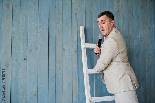 Handsome man in beige suit and pink shirt with microphone against ladder background on studio. Funny face of toastmaster and showman.