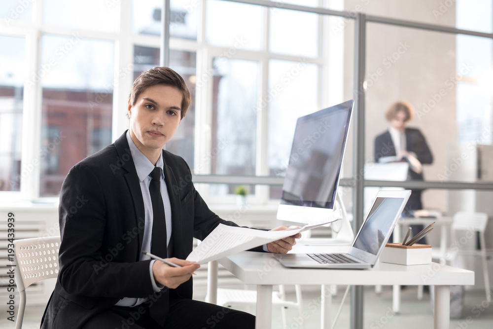 Serious and confident businessman with papers looking at camera by workplace while looking through new contracts Serious and confident businessman with papers looking at camera by workplace while looking through new contracts