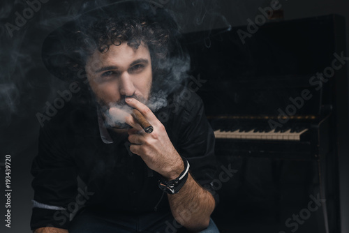 handsome musician smoking cigar in front of piano on black