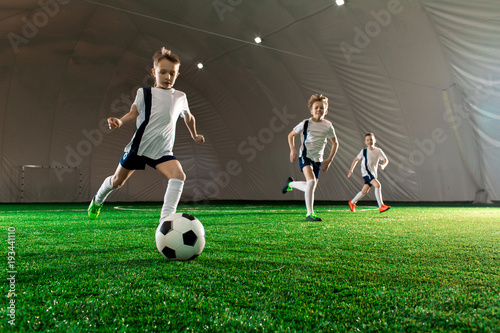 Three boys in uniform running after ball down green field while training indoors