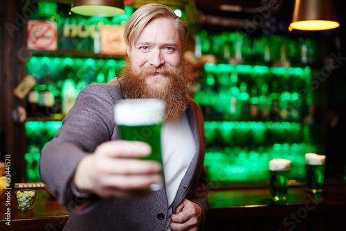 Cheerful bearded man looking at camera while cheering up with glass of foaming beer