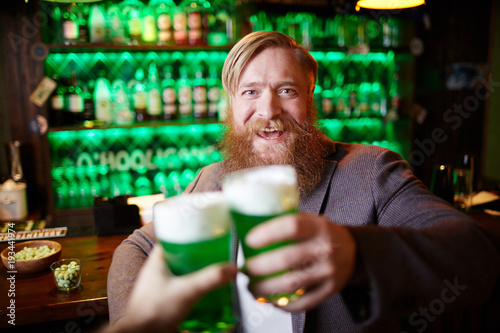 Excited bearded man with glass of foaming beer clinking with his friend in pub